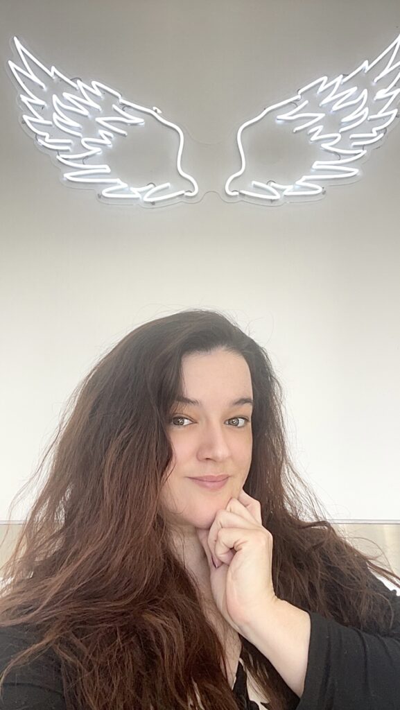 Long haired brunette woman Elizabeth Best in front of neon angel wings sign