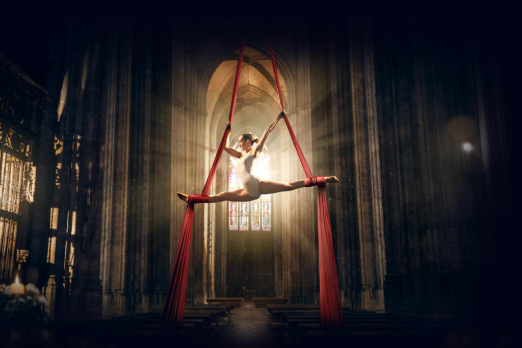 A woman is doing the splits at the top of a set of red silks in a church for Eternity for Brisbane Festival.