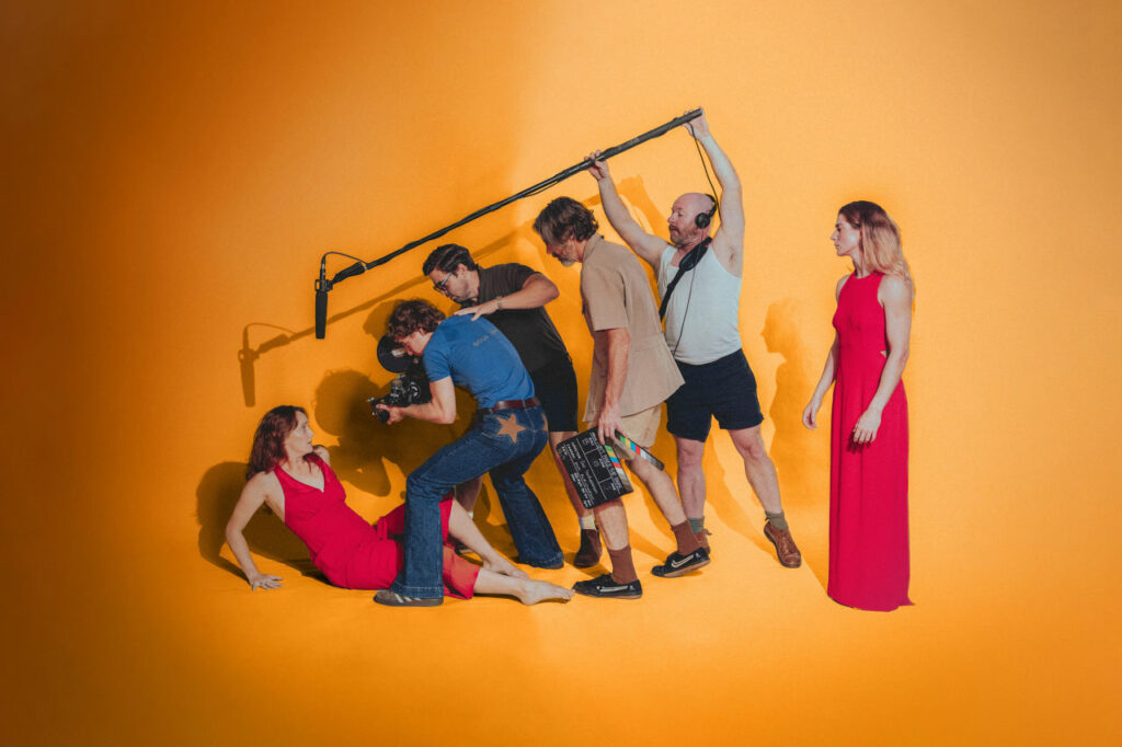 An actress in a red dress is being surrounded by people in the film industry from camera people to boom mike operators to director for Stunt Double Brisbane Theatre.