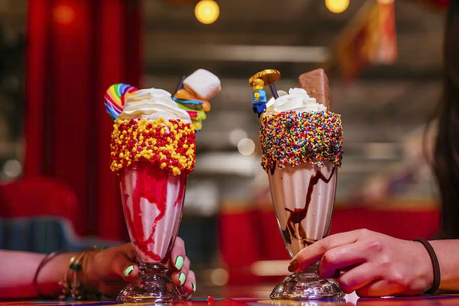 Two cocktails in sundae glasses with sprinkles all over the rims, whipped cream on top and chocolate bars and lolly pops sticking out the top on a table at Archie Brothers Cirque Electriq. 