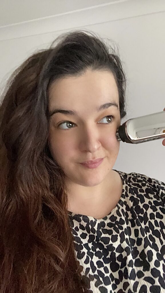 Brunette woman with long hair holds CurrentBody Skin Radio Frequency Device up to her cheek. Cheeks are slightly red. 