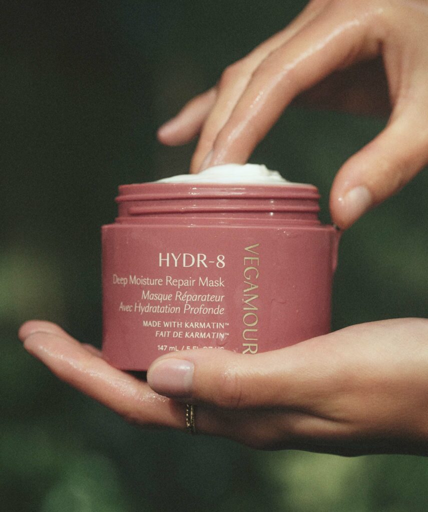 A hand holds a tub of Hydr-8 by Vegamour while the other hand scoops out product.