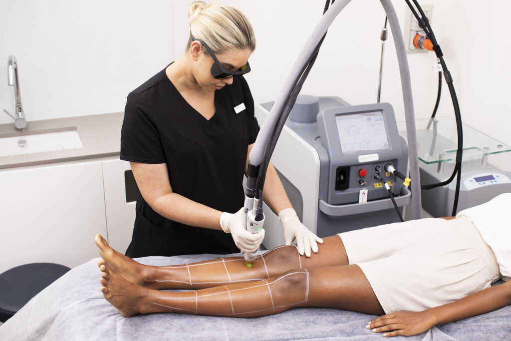 A woman dressed in black scrubs administers laser hair removal on a woman lying on a bed. 