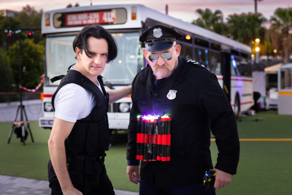 Two men stand in front of a bus looking menacing for Speed: The Movie The Play. 