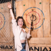 Girl with short brown hair and glasses standing in front of a target, holding an axe in one hand, cheering. Maniax is written on the wall.