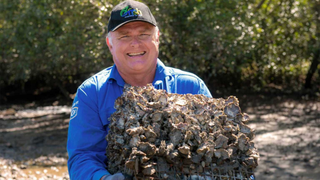 A man with grey hair smiles while holding a rock covered in oysters.