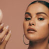 Selena Gomez with a Rare Beauty compact in her hand and a makeup brush touching her face in the other
