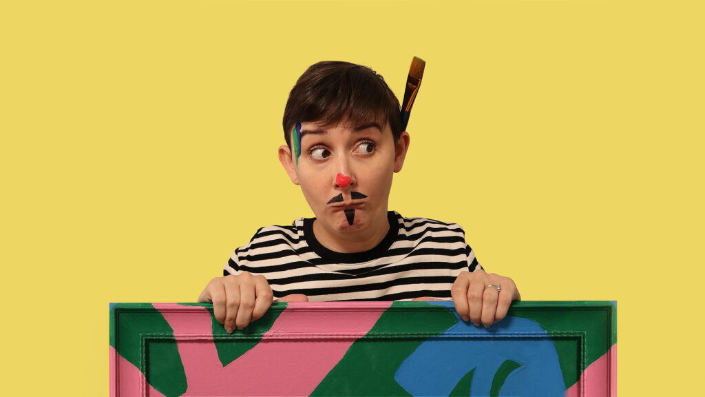 A woman with a paint brush behind one ear and a painted on red nose and mustache poses behind an art canvas for the show Artiste. 