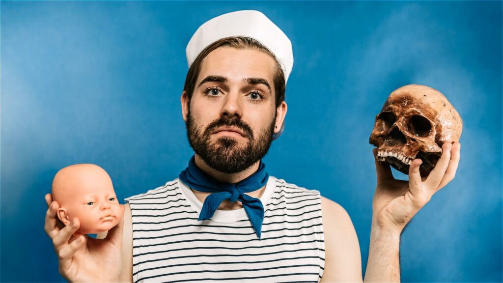 A man in a sailor suit poses on a blue background with a doll head in one hand and a skull in the other for the show Spunk Daddy. 