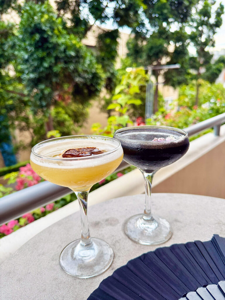 Two cocktails sit on a table, in front of a balcony rail and plants.