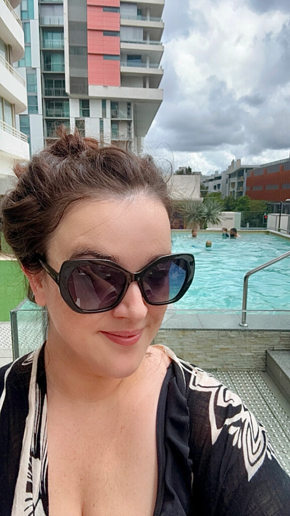 A girl with brown hair and big sunglasses in a black bathing suit smiles at the camera. There is a pool in the background. 