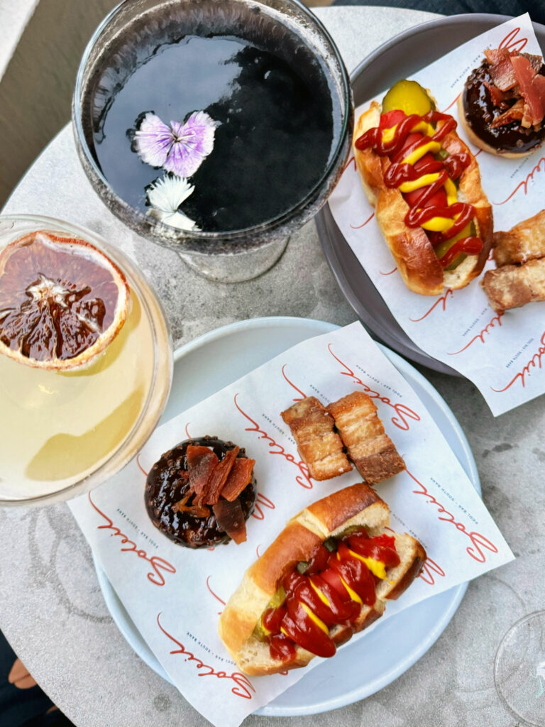 Top down photo of two cocktails and a grazing platter of hot dogs, donuts and pork belly. 