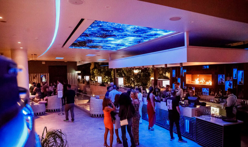 People mill around a lively bar at night time, the bar has an LED screen on the ceiling projecting starlight.