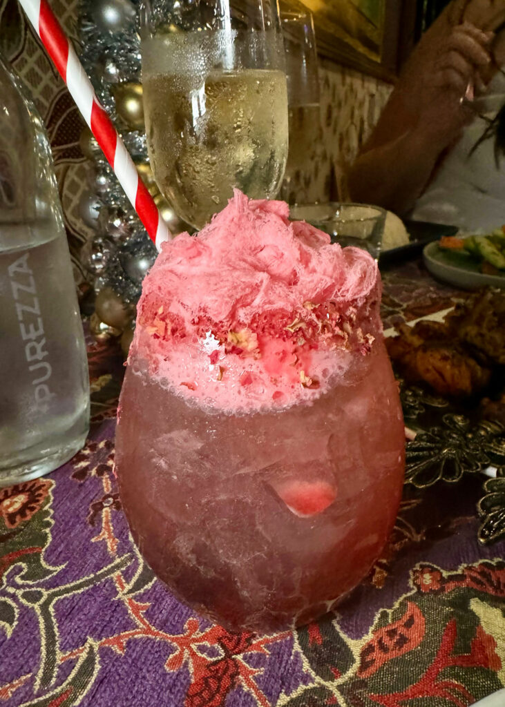 A pink cocktail with fairy floss and rose petals on top with a straw. 