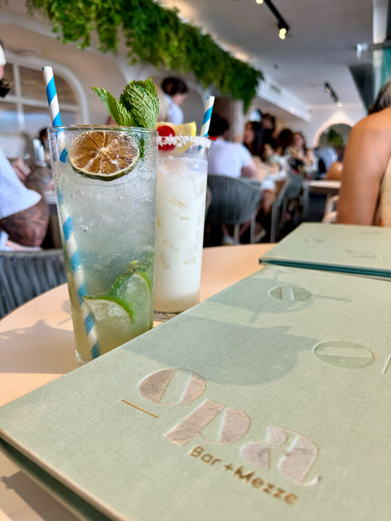An Opa menu sits in the foreground with a mojito and pina colada behind it. 