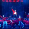 Five men dressed in red jackets pose on top of a red car under stage lights in the musical Grease