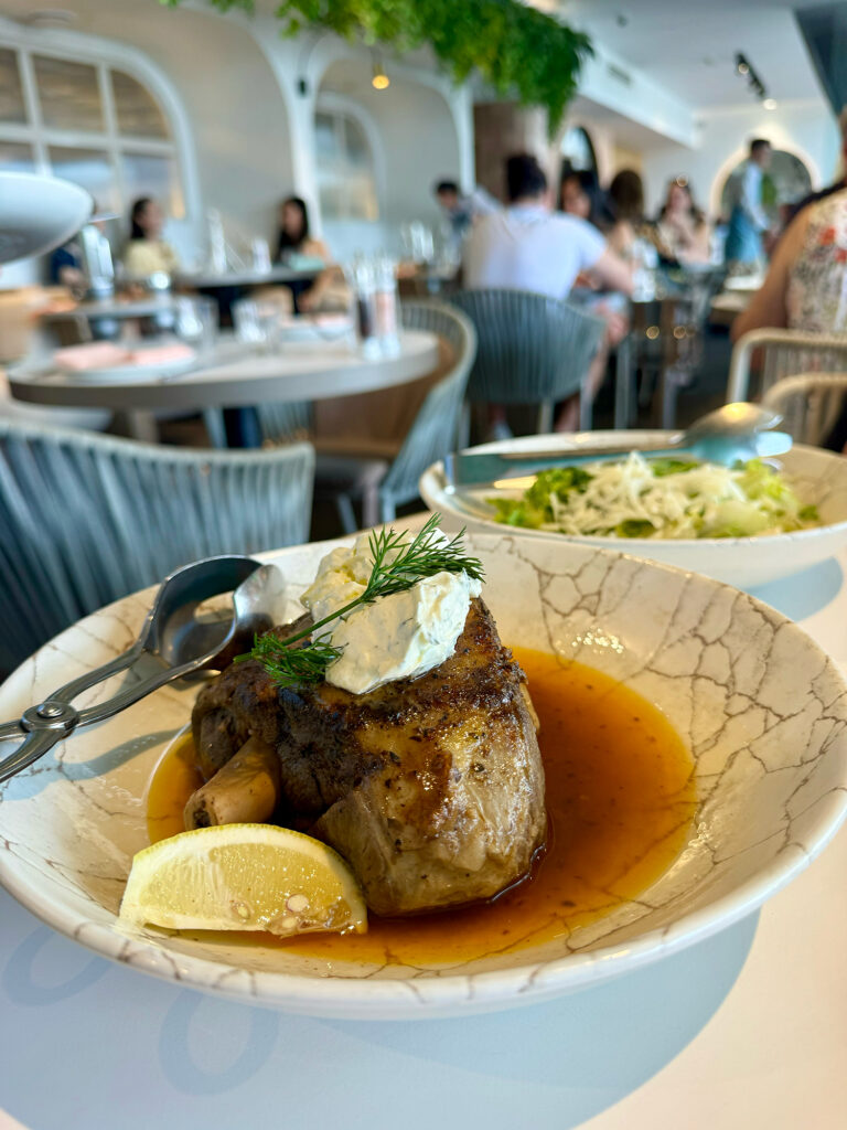 A lamb shoulder sits on a marbled bowl, garnished with yoghurt and a slice of lemon, with a lettuce salad in the background on a white table. 