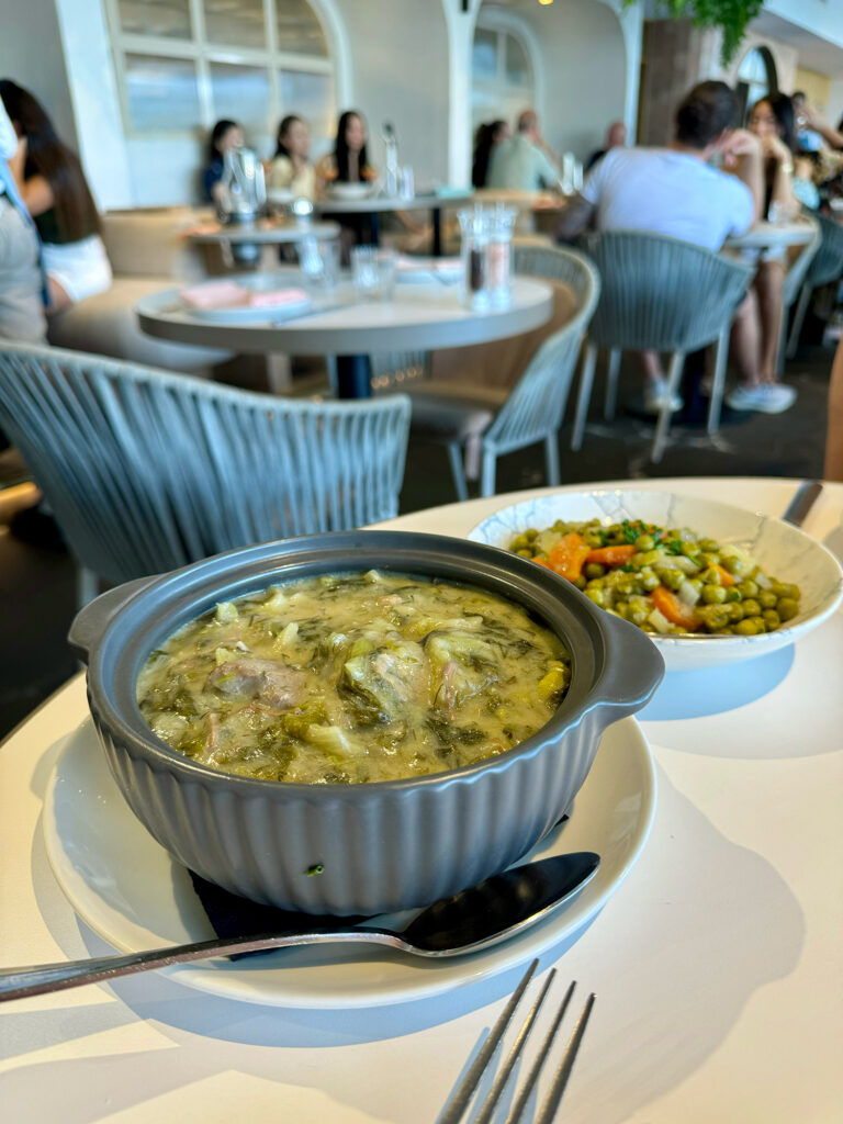 A bowl of lamb fricassee sits in the foreground with a spoon beside it, with a bowl of peas and carrots behind it on a white table. 