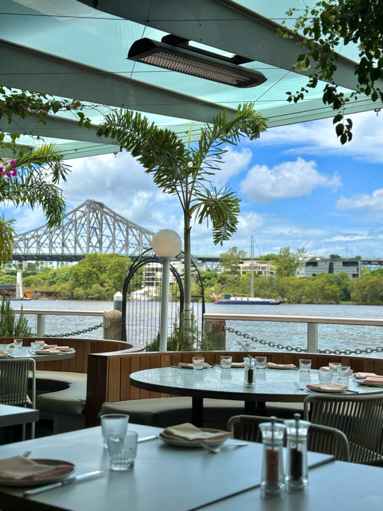 Restaurant tables sit beside the Brisbane river on a sunny day, with the Story Bridge visible in the background.