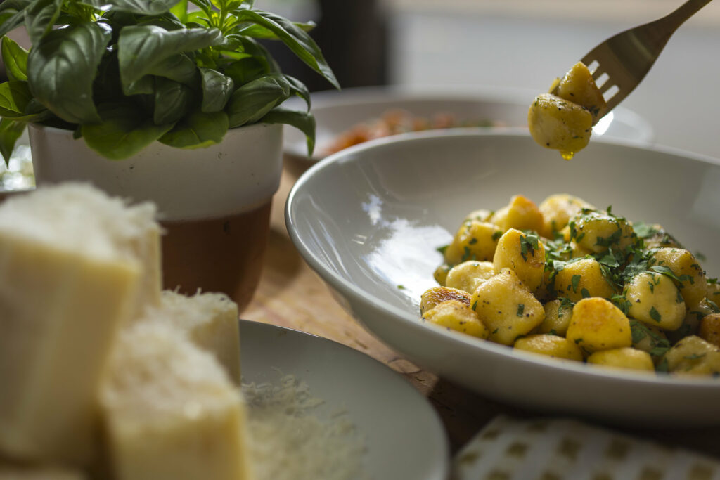 A fork is picking up two gnocchi pieces off a plate of plentiful gnocchi. A plate of basil and parmesan cheese is off to the side.