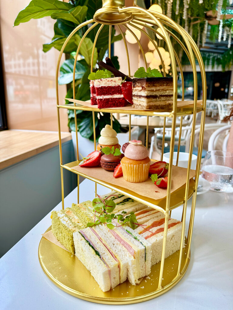 A golden half bird cage with three tiers, with cake on the top tier, cupcakes and macarons, strawberries on the second tier and a bottom tier of ribbon sandwiches.