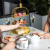 two women sit across the table from one another, one is reaching for a cake on a three tier high tea stand.