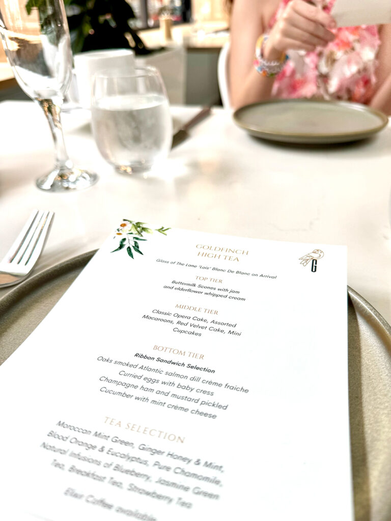 A Goldfinch high tea menu sits on a grey plate, with a glass of champagne above it on a white table