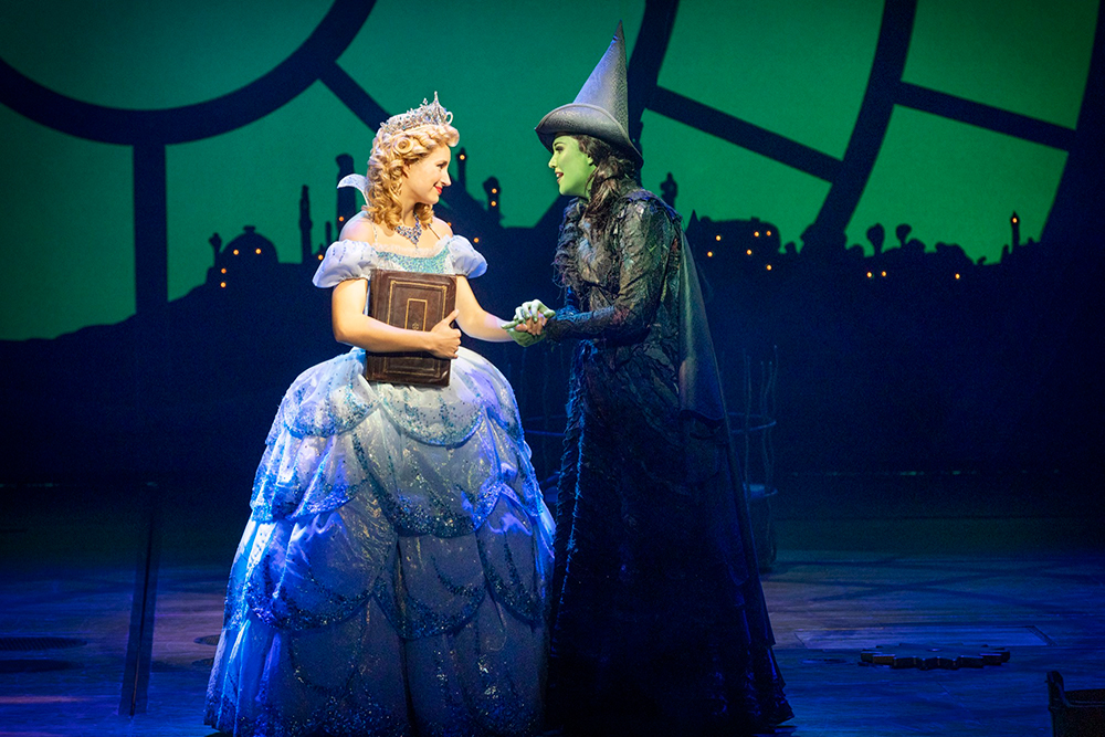Glinda the good witch holds hands with Elphaba in on a green background in Wicked the Musical