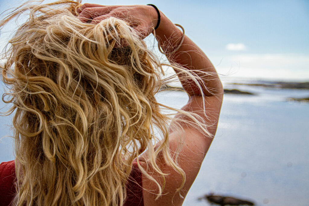 A blonde woman with wavy hair is seen putting her hands in her hair from behind as she stands on a beach.