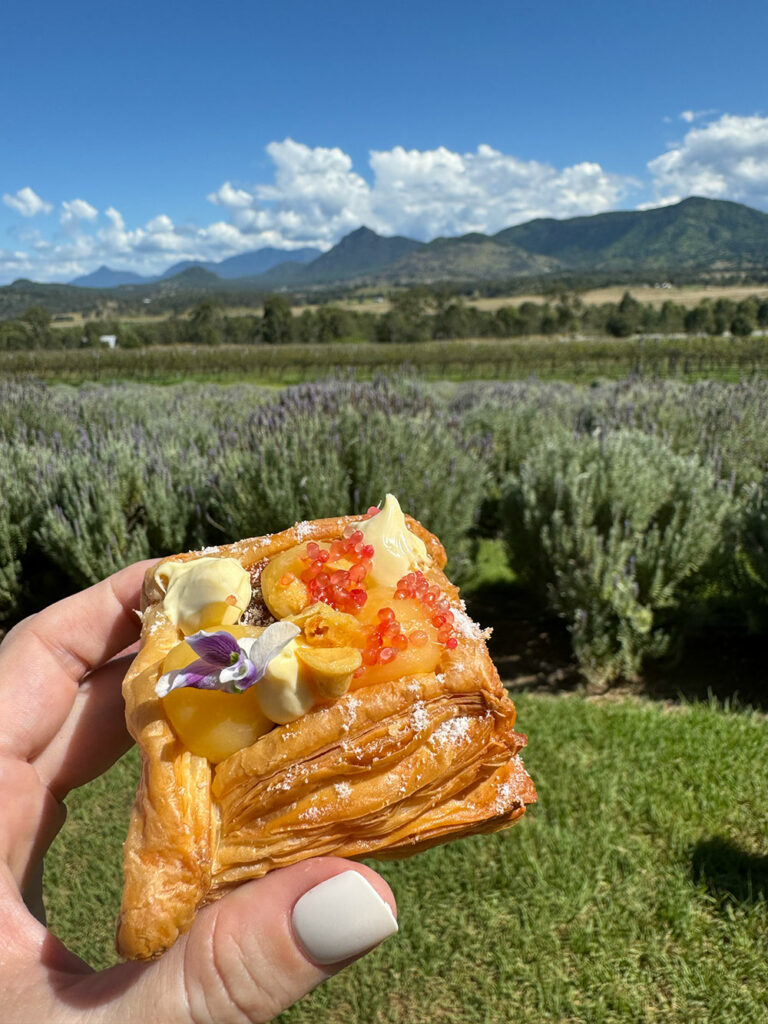 A hand with white fingernails holds up a danish filled with creme fraiche, topped with lime curd, fingerlime and an edible flower, with a lavender farm in the background.