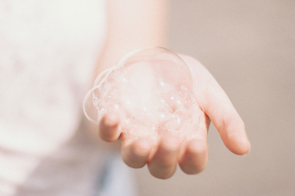 A white woman's hand is outstretched to the camera, and is filled with shampoo bubbles.