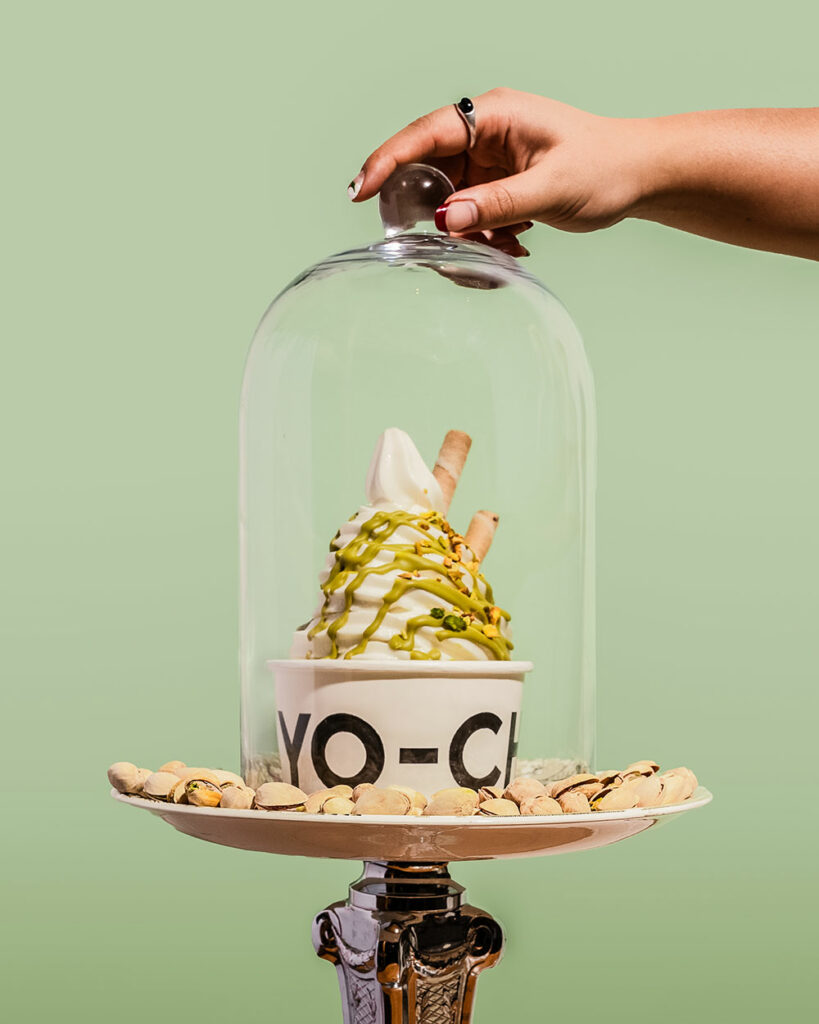 A full tub of yo-chi frozen yoghurt covered in Pistachio Papi spread sits underneath a glass cloche with a hand poised to lift it. 