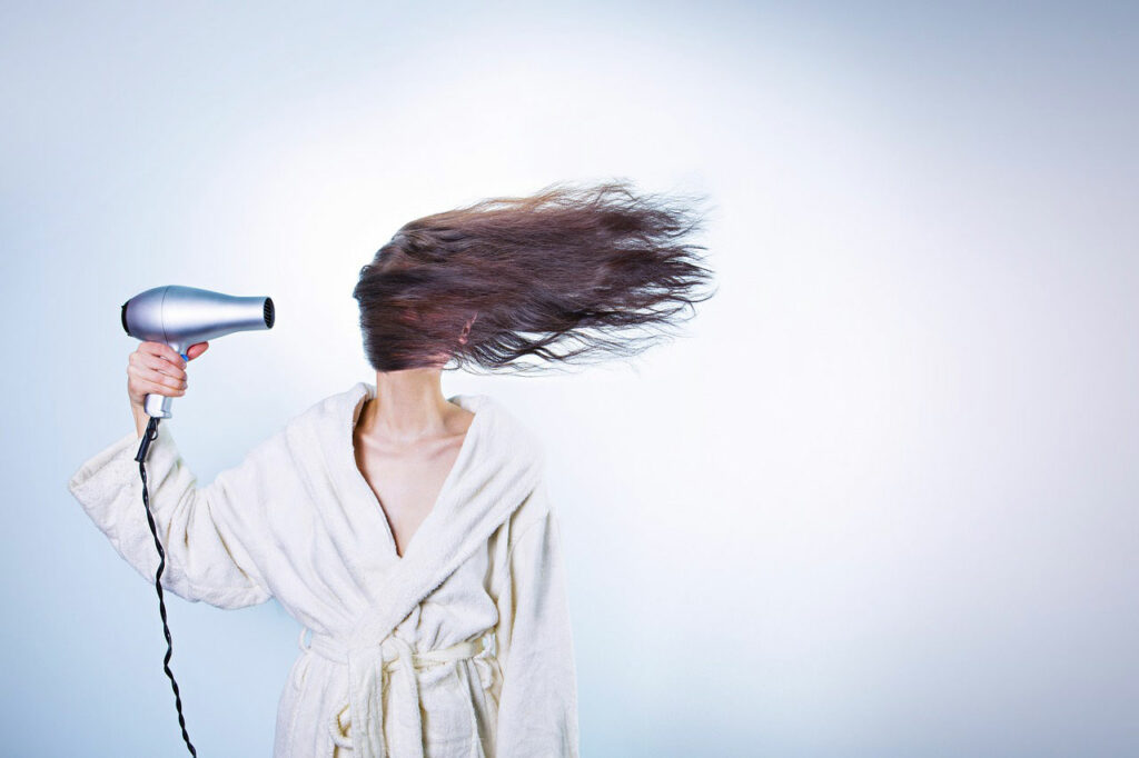 A brunette woman holds a hairdryer pointing at her head, with her hair being blown in front of her face