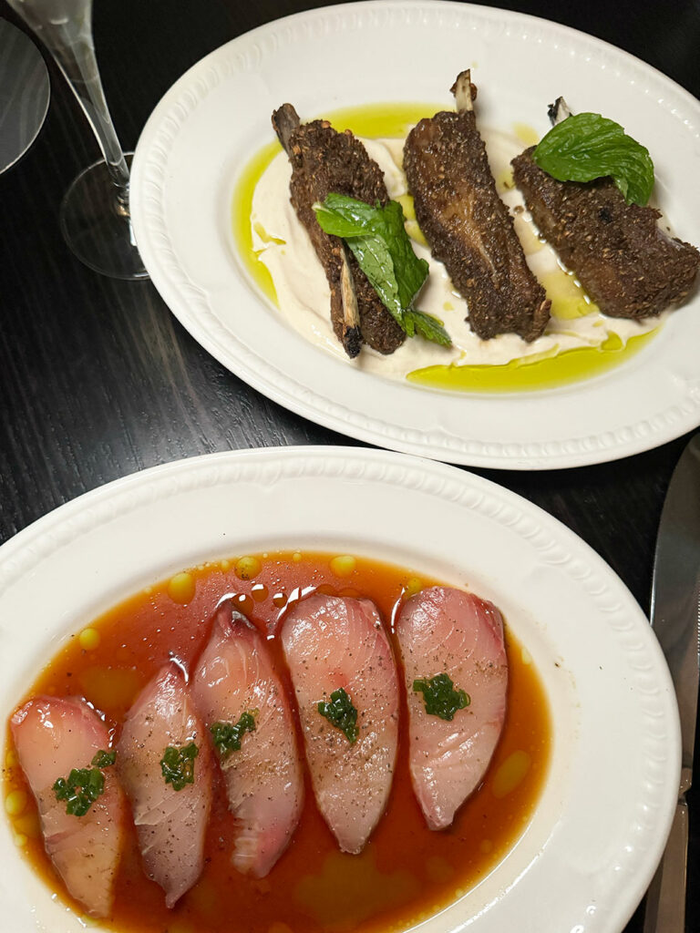 A plate of lamb ribs and a plate of kingfish in red sauce sits on a black table. 