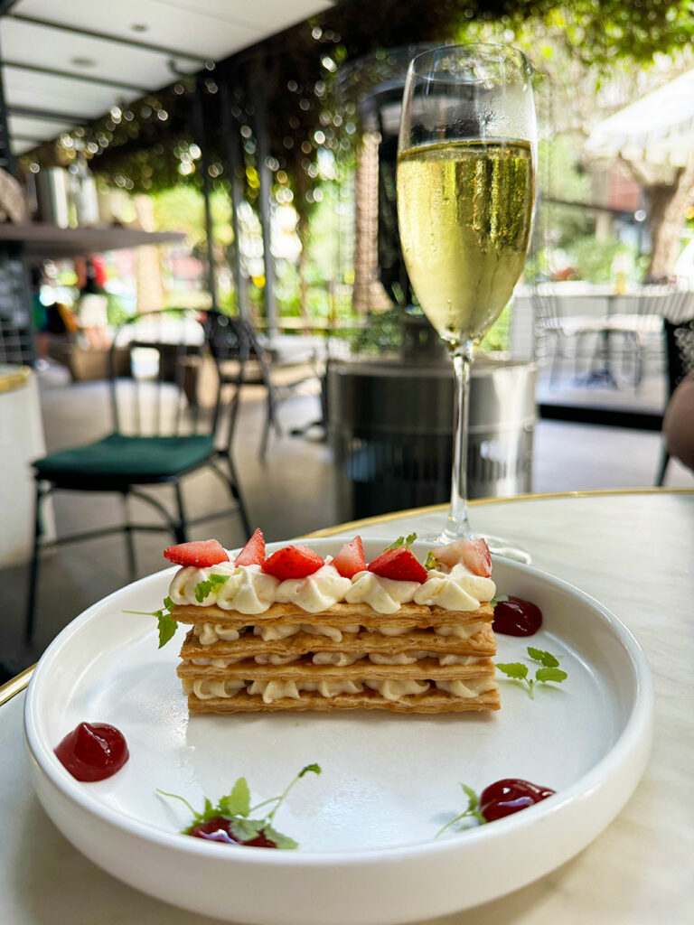 A mille-feuille topped with strawberries with a glass of champagne behind it, greenery of the restaurant behind it. 