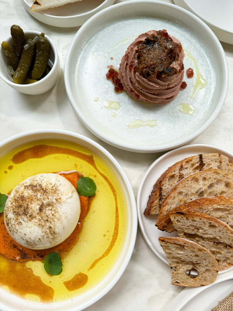 Three plates on a white table, one with burrata, one with pate and one with sourdough bread. A small bowl of cornichons sits next to it.