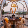 A see through igloo tent sits on the brisbane river, with a cosy table and chairs setup inside