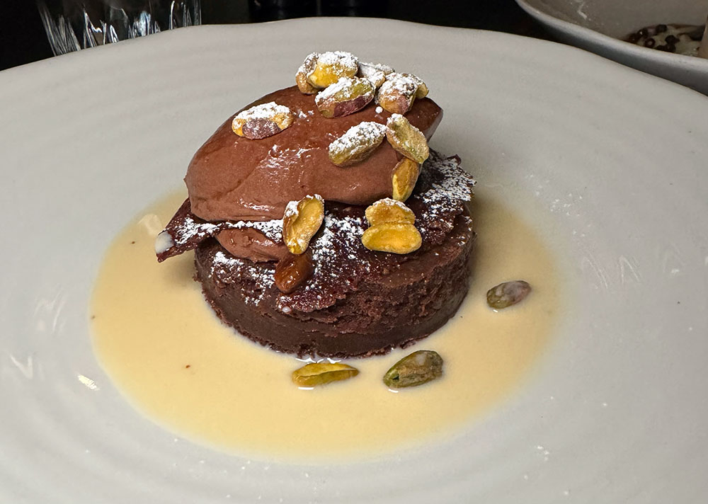 A round slice of chocolate cake topped with mousse, pistachios and creme anglaise.