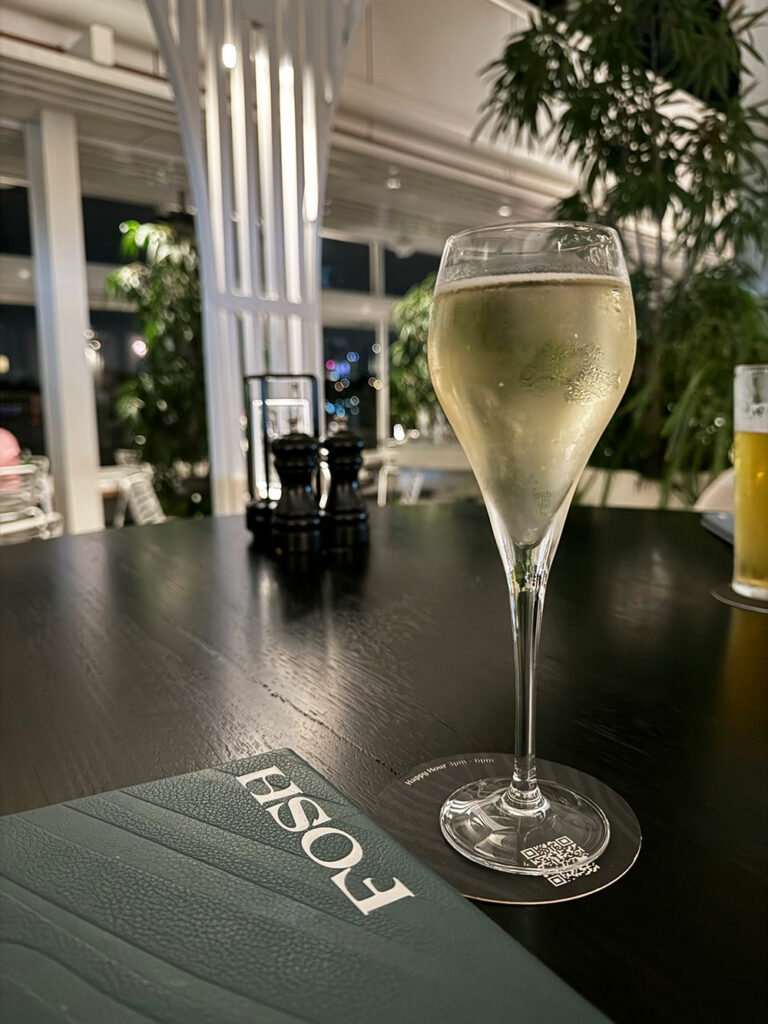 A glass of white wine and a FOSH menu sits on a black table, surrounded by greenery.