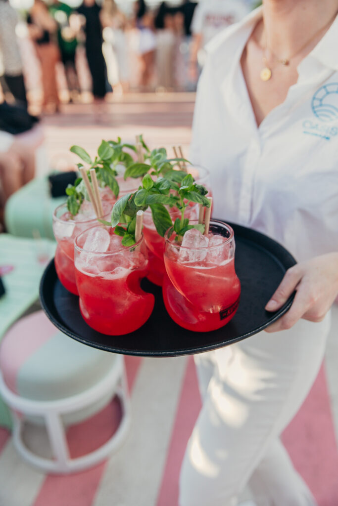 A woman holds a tray of red cocktails garnished with mint springs, with pink and white striped carpet of OASIS Brisbane visible below her feet. 