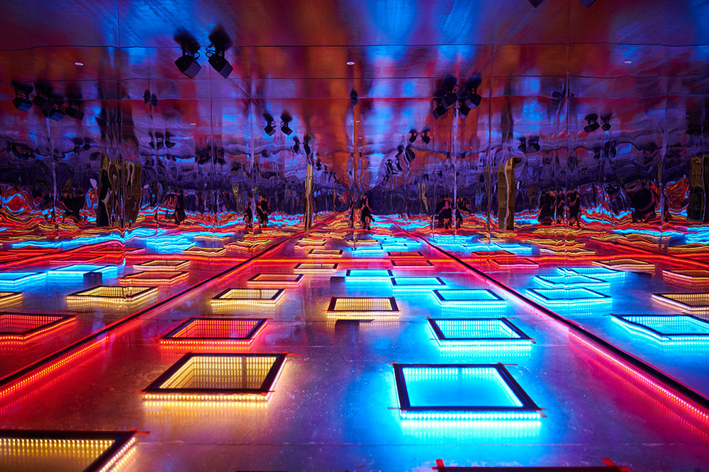 A room with lit up squares of different colours on the floor surrounded by mirrors. 