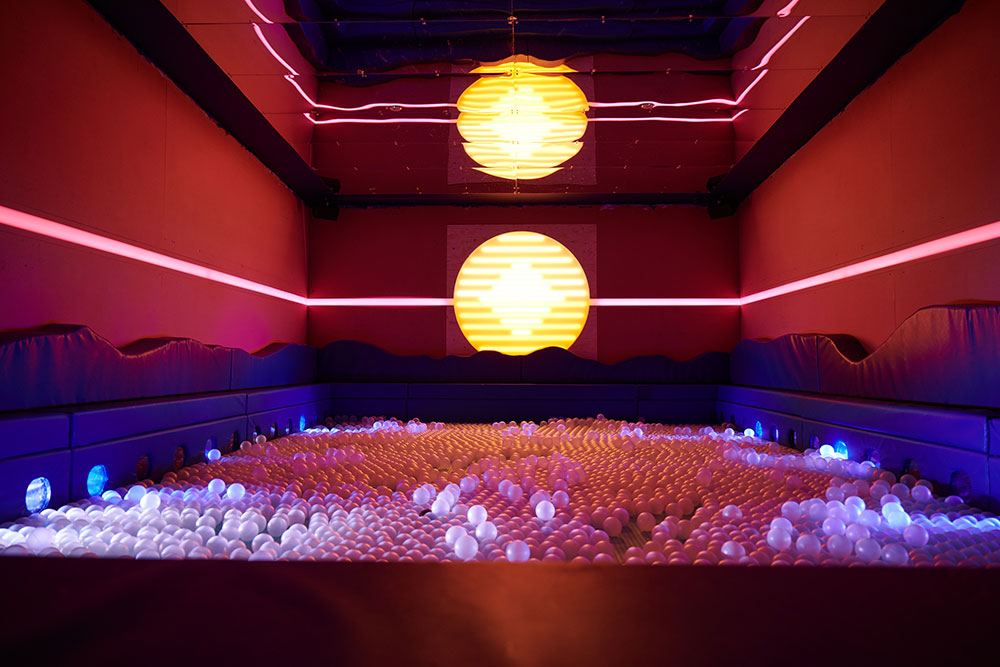 A ball pit filled with white balls is surrounded by a miami beach mural with a neon sun.