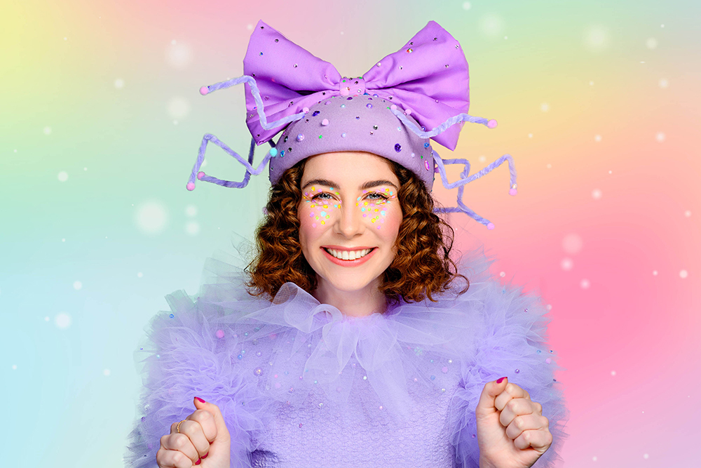 A woman with curly red brown hair wears a lilac tulle top and a hat with purple sparkles and a big bow and some pipe cleaner spider legs. 