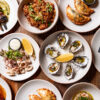 A selection of Argentinian dishes sits on a table, including oysters, calamari, empanadas, lamb skewers and queso cheese.