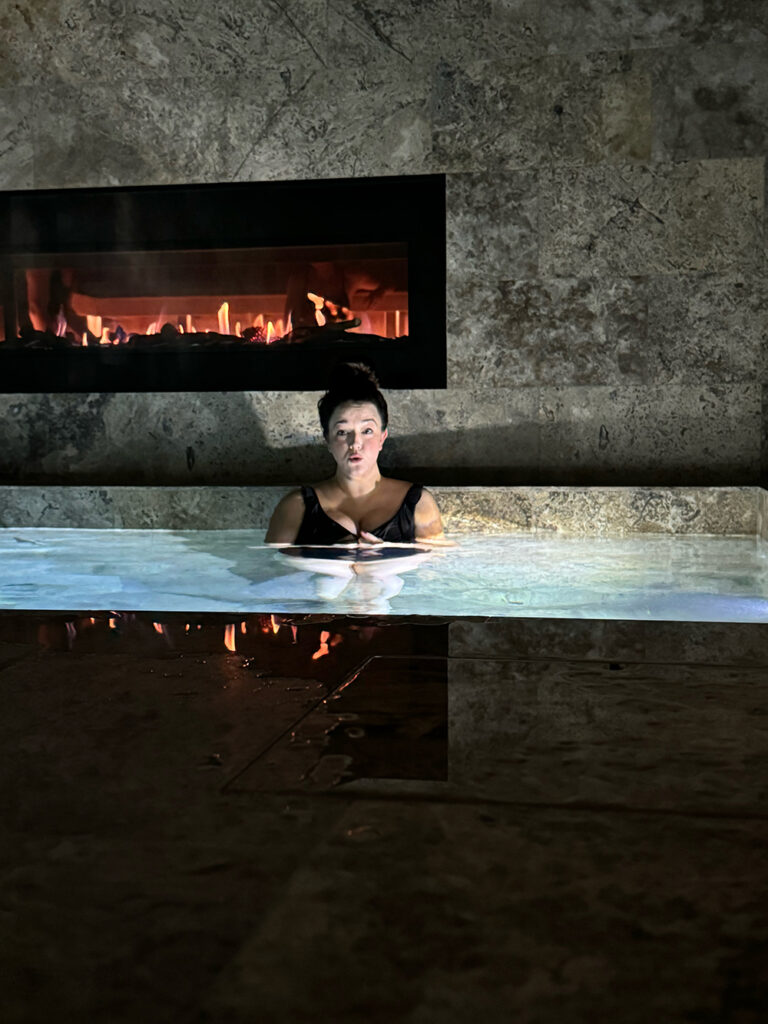 A brunette woman with a messy bun stands in a pool with water up to her chest with a surprised look on her face. 