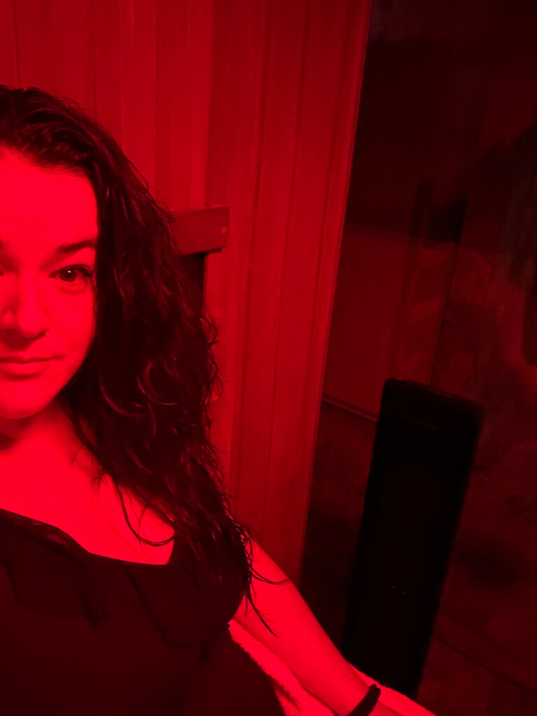 A brunette woman in a black bathing suit is in an infrared sauna and there is a red light tint over her whole body. 