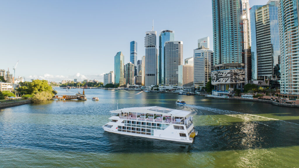 A three level party boat cruises on the brisbane river with buildings in the background along the river bank