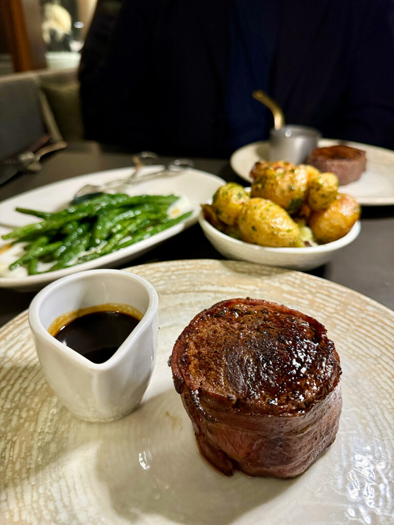 A filet mignon steak wrapped in bacon at the foreground, with a plate of green beans and a bowl of kipfler potatoes in the back. 