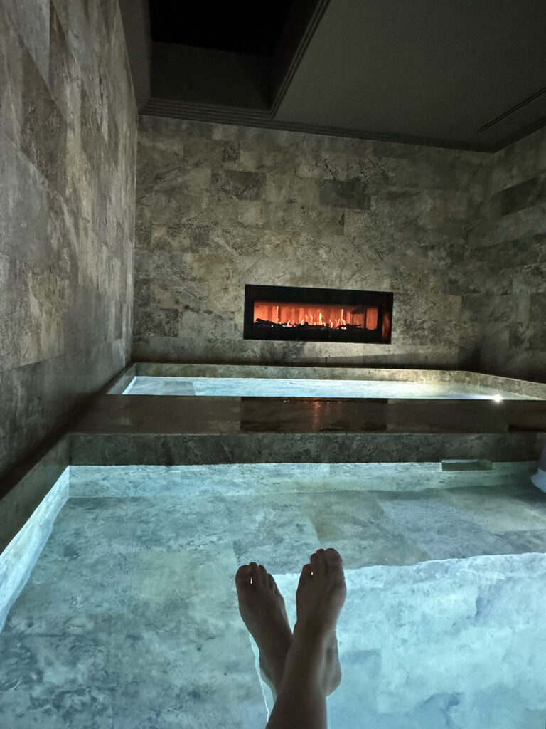 A pair of feet crossed at the ankles float in a pool, facing a fireplace. 