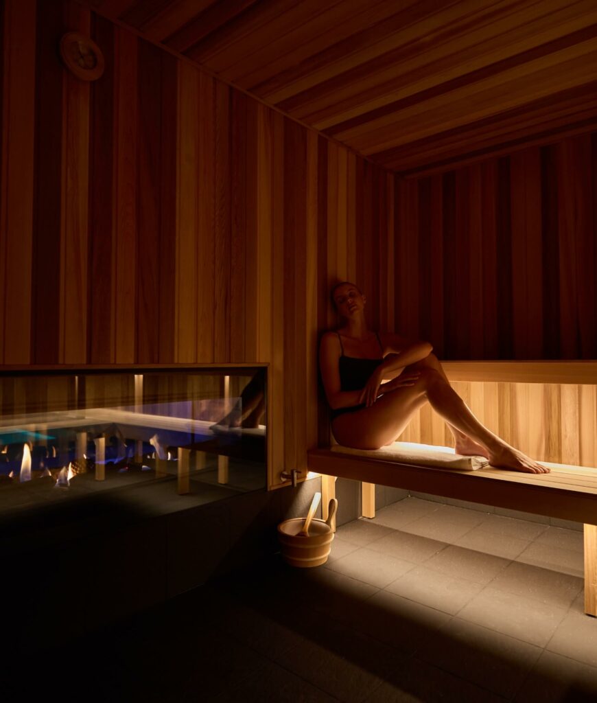 A woman sits with her legs stretched out on a wooden sauna ledge, and behind a glass panel there is a fireplace to her left.